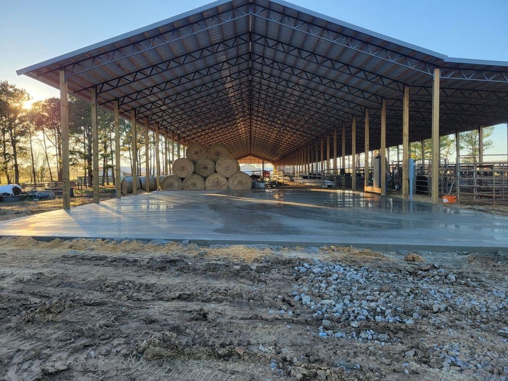Large agricultural barn interior with hay bales, concrete floor, and sunlight filtering through.