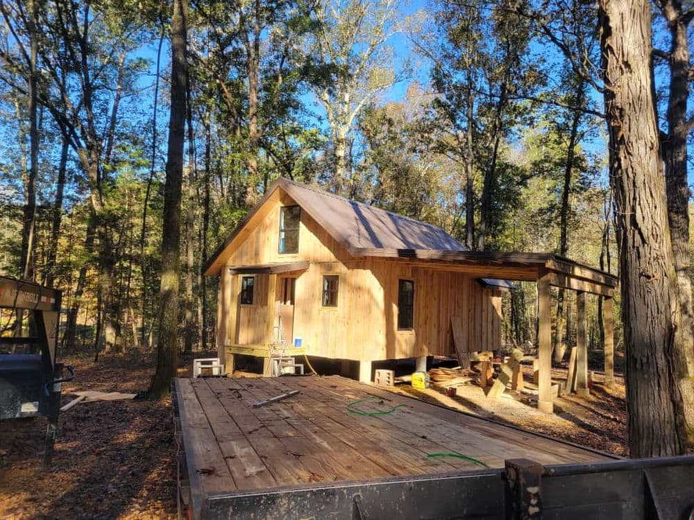 Wooden cabin under construction in a wooded area, surrounded by trees and clear blue sky.